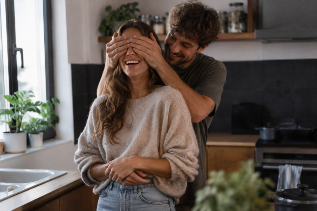 "Foto autêntica e espontânea de um casal na cozinha. O homem está vendando os olhos da mulher com as mãos por trás, guiando-a para uma surpresa. Ela está dando uma risada gostosa, com uma expressão de expectativa. Ambiente caseiro, iluminado com luz natural entrando pela janela. Fotografia com textura suave."