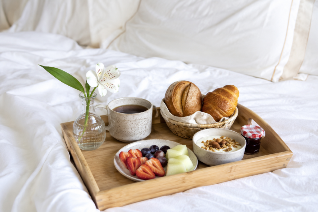 Uma bandeja de café da manhã simples e charmosa em cima de uma cama arrumada com lençóis brancos, com pães, frutas e um pequeno vaso com uma flor. Luz natural." presente de dia dos namorados